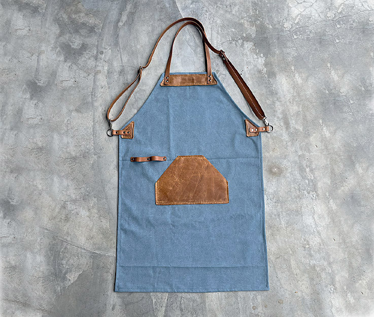 A sky blue canvas apron with tan leather detail showing the new adjustable crossover straps featured against a concrete screeded floor.