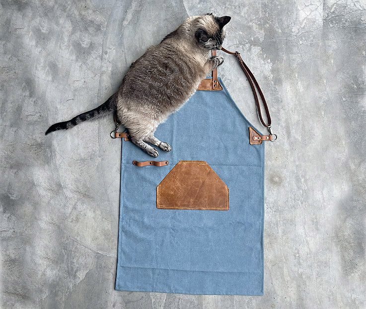 A sky blue canvas apron with tan leather detail with Cyrus the garage cat featured against a concrete screeded floor.