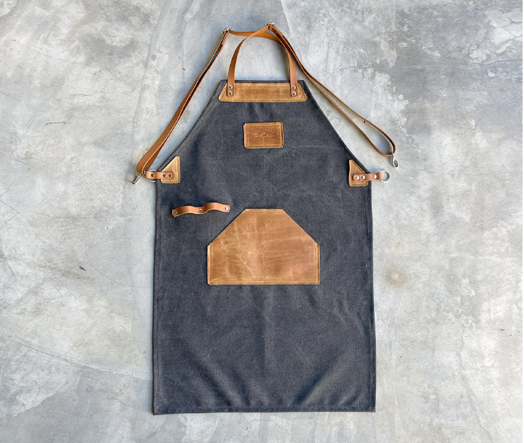 A washed black canvas apron with tan leather detail and straps.  The updated apron design has crossover straps. Apron featured against a concrete screeded floor.