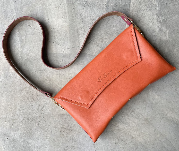 A coral coloured leather clutch sized bag with rose pink detail, stunning curved lines, and studs along the base.