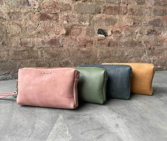 A collection of luxury leather toiletry bag in an assortment of beautiful colours against a rough brick wall and concrete floor.