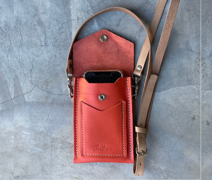 The Smithco Essential mini leather bag in coral with a phone to compare the size. Featured with a detachable and adjustable leather strap set against a concrete screed floor.