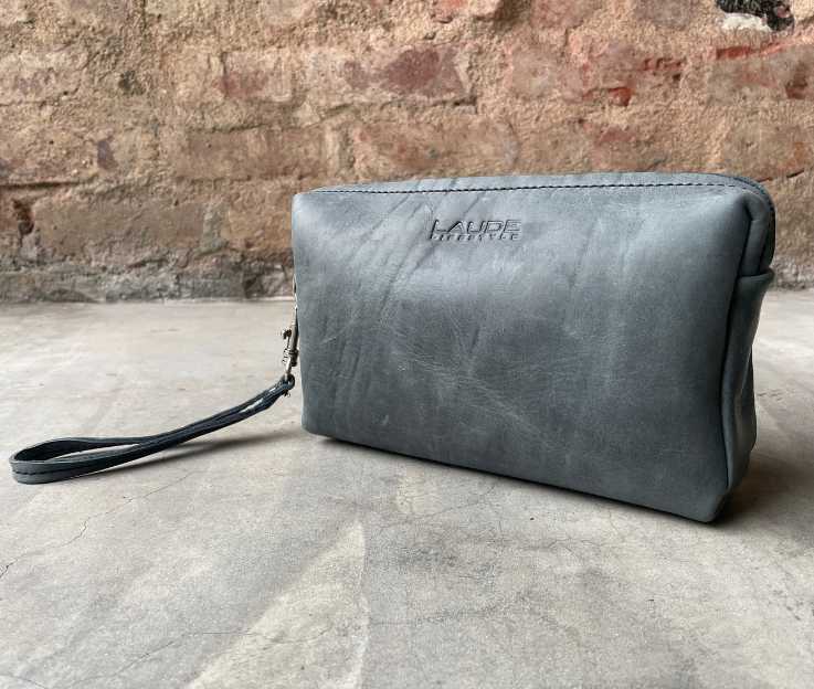 A luxury leather toiletry bag in denim blue against a rough brick wall and concrete floor.