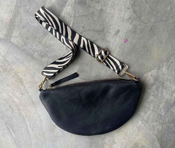 A black half-moon bag with a matching decorative adjustable crossbody strap set against a concrete screeded floor.