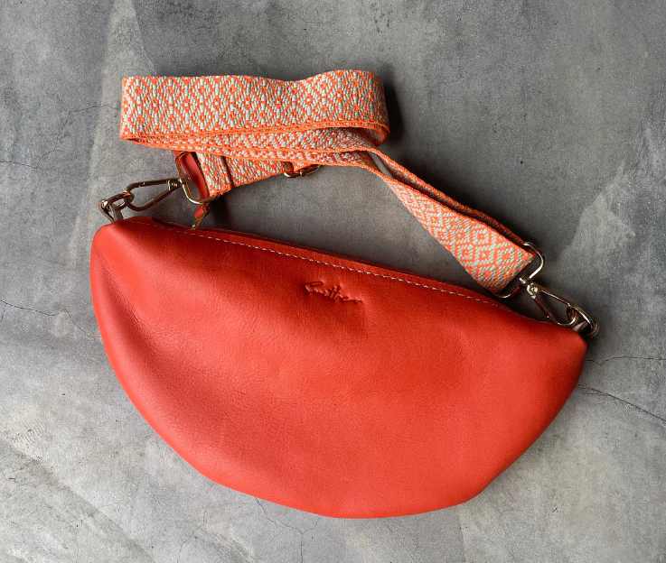 A coral coloured half-moon bag with a matching decorative adjustable crossbody strap set against a concrete screeded floor.