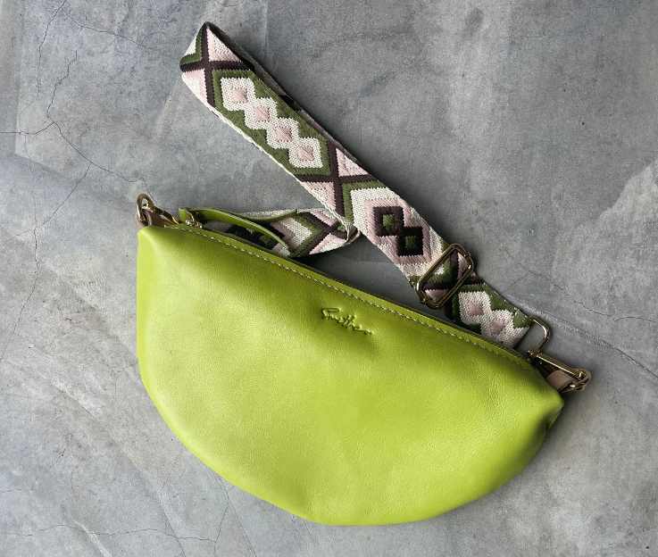 A lime coloured half-moon bag with a matching decorative adjustable crossbody strap set against a concrete screeded floor.