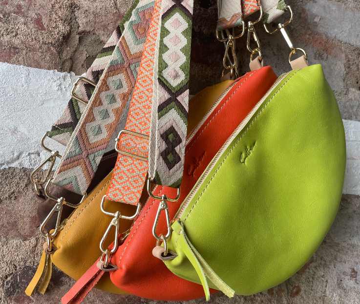 A collection of half-moon bags in vibrant colours hanging against a rough red brick wall.