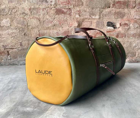 Angled view of a full grain leather duffel bag in a dark forest green with Ochre coloured panels and brown details pictured against a concrete screed floor and rough red brick wall. This bag features corporate branding.