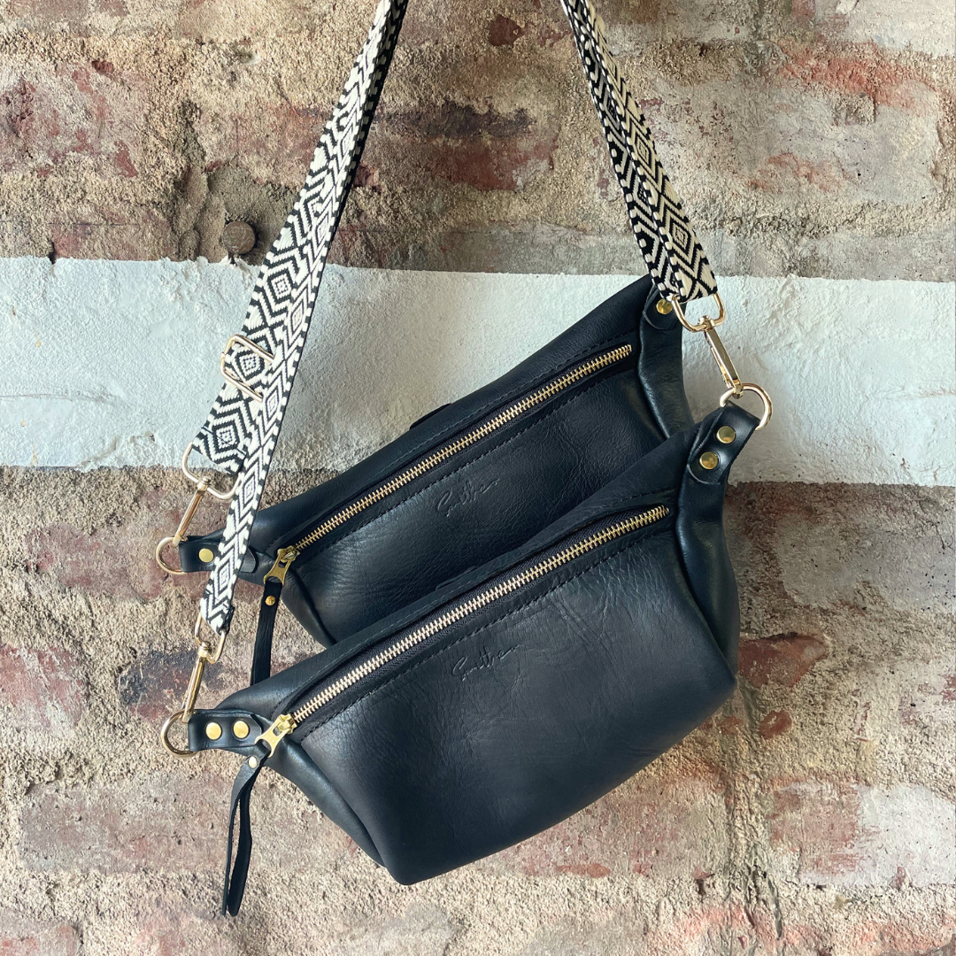 The black moon sling bag displayed from the front against a rough red brick wall.