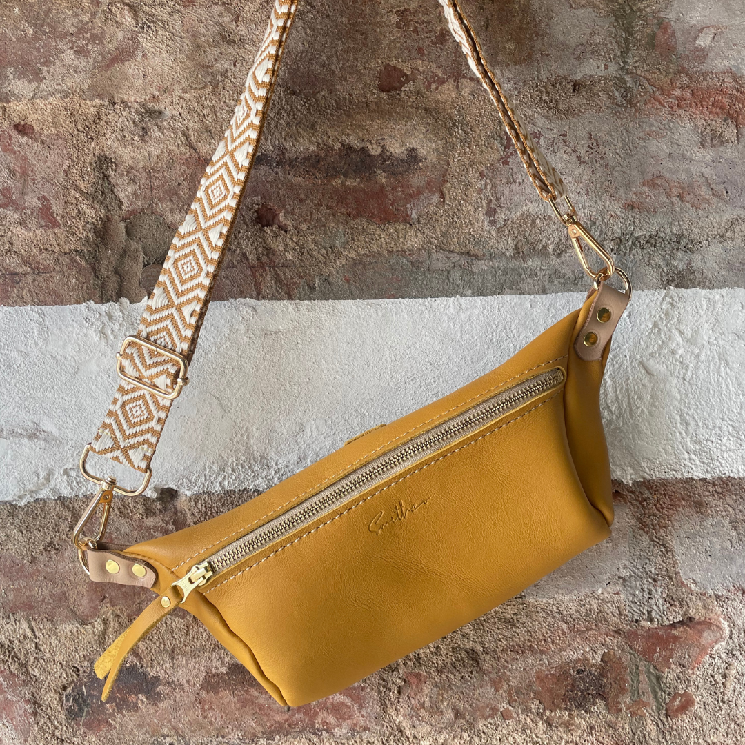 The ochre coloured moon sling bag displayed from the front against a rough red brick wall.