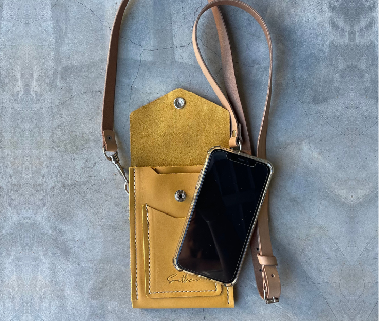 The Smithco Essential mini leather bag in ochre with a phone to compare the size. Featured with a detachable and adjustable leather strap set against a concrete screed floor.