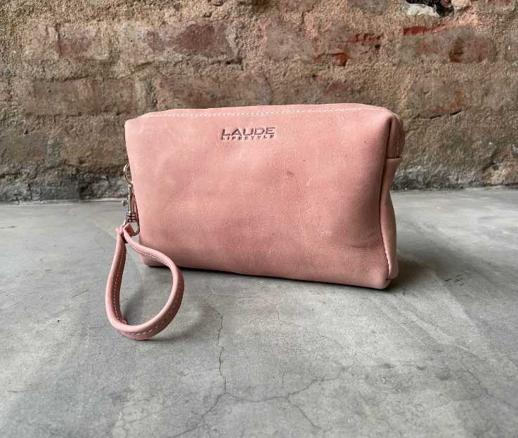 A luxury leather toiletry bag in dusty pink against a rough brick wall and concrete floor.