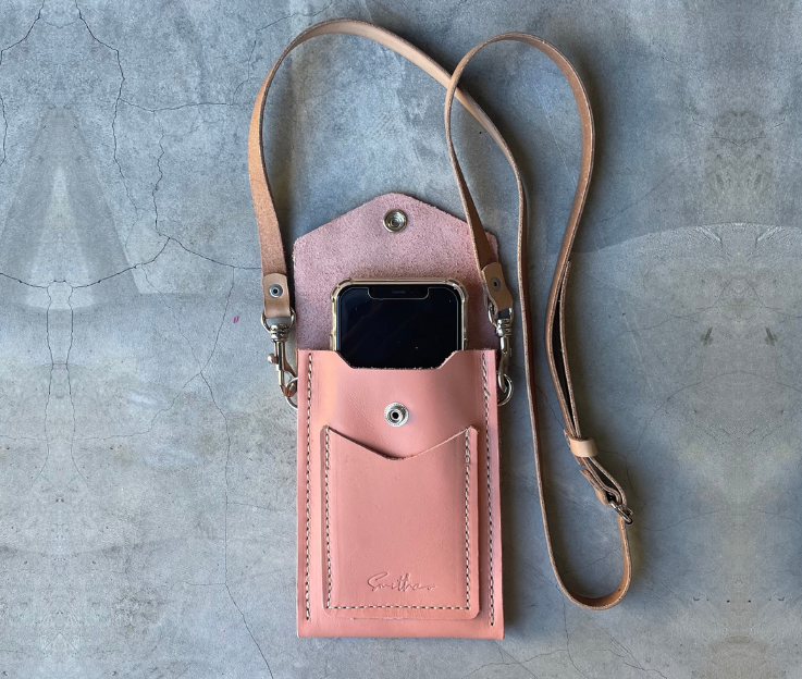 The Smithco Essential mini leather bag in rose pink with a phone to compare the size. Featured with a detachable and adjustable leather strap set against a concrete screed floor.