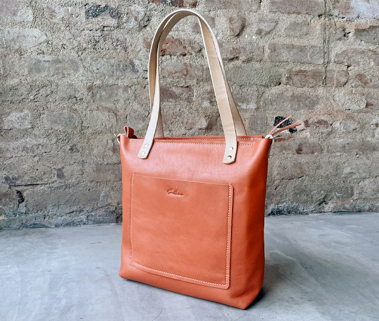 Sassy Shopper steals the spotlight against a sleek concrete screen wall, framed by a backdrop of rustic red brick. The vibrant leather bag in Coral pops with personality, adding a bold touch to the urban-chic scene. #SmithcoSassy #UrbanStyle"