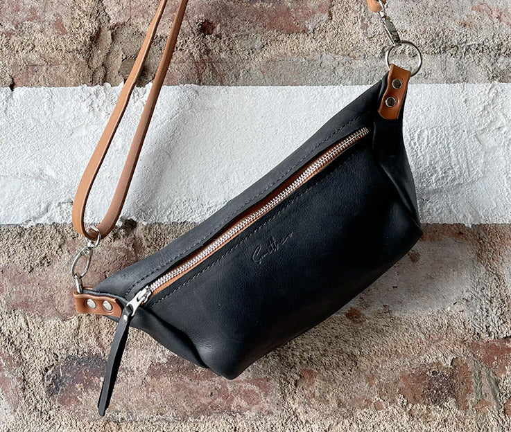 The black and amber coloured moon sling bag displayed from the front against a rough red brick wall. 