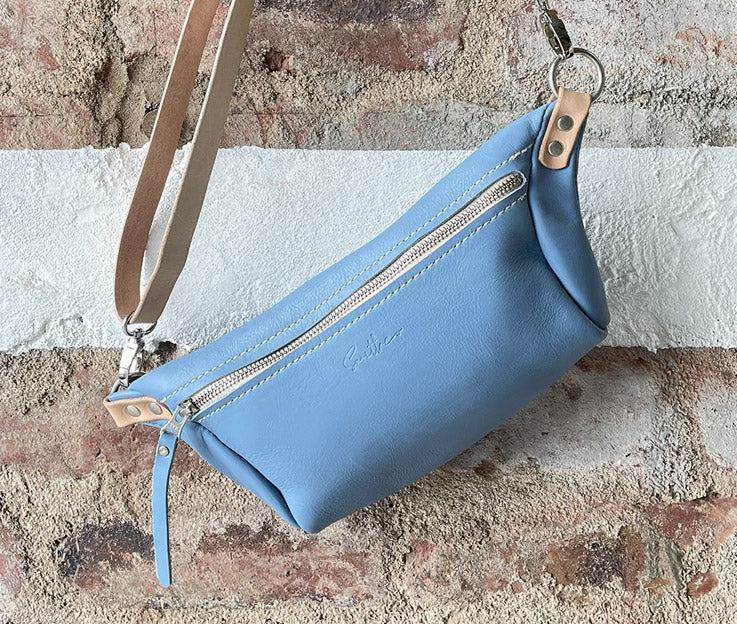 The sky blue and nude coloured detail moon sling bag displayed from the front against a rough red brick wall. 