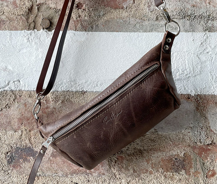 The diesel brown moon sling bag displayed from the front against a rough red brick wall.