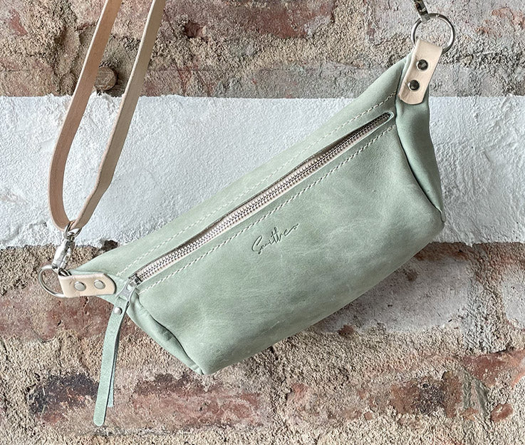 The sage green with nude coloured detail moon sling bag displayed from the front against a rough red brick wall. 