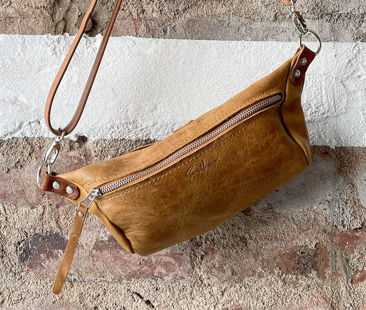 The diesel toffee coloured moon sling bag displayed from the front against a rough red brick wall. 