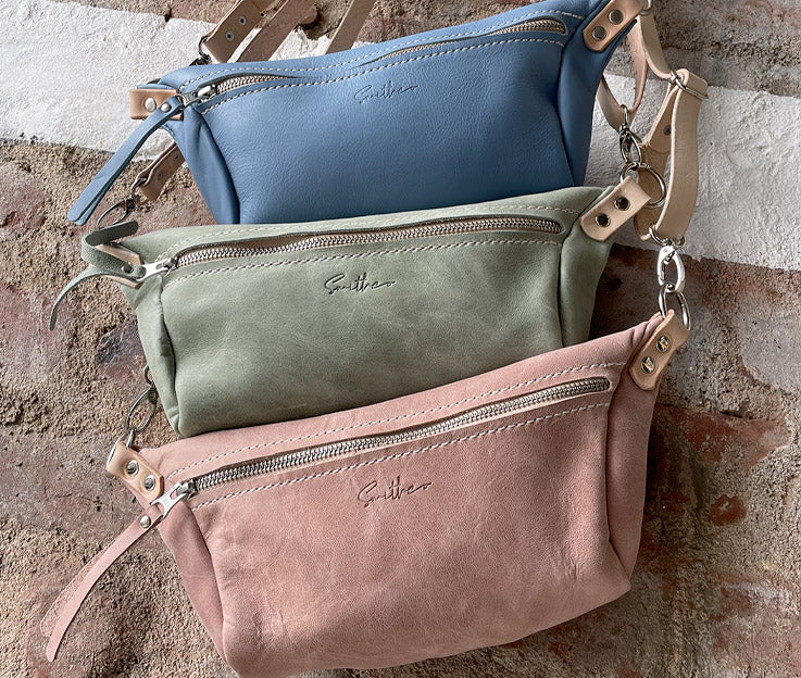 Three moon sling bags hanging against a rough red brick wall. Colours are sky blue, sage green and dusty pink, all with nude coloured detail.