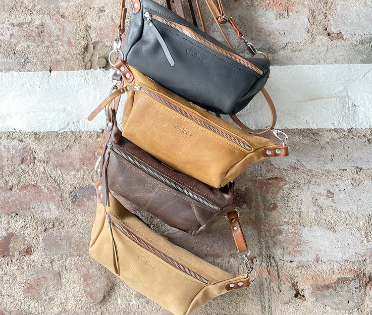 Four moon sling bags hanging against a rough red brick wall. Colours are black and amber, diesel toffee, diesel brown, and fudge.