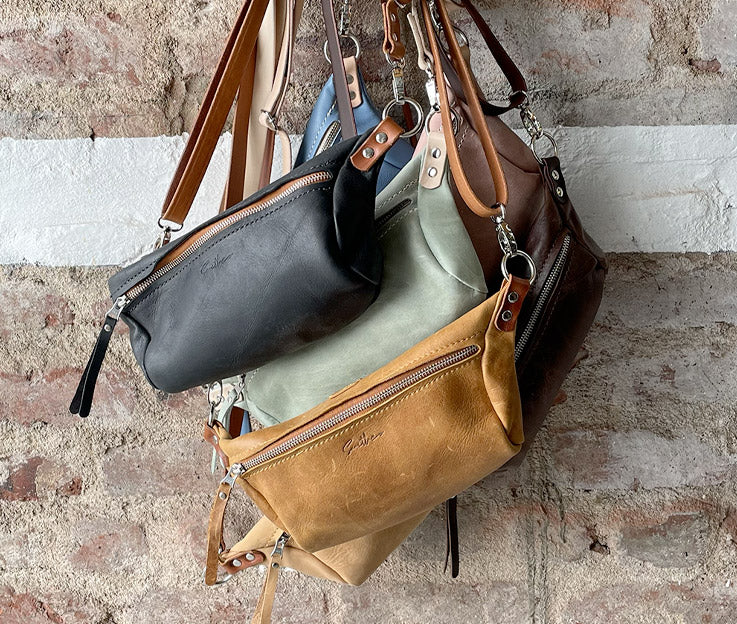 A collection of moon sling bags in various colours hanging against a rough red brick wall.