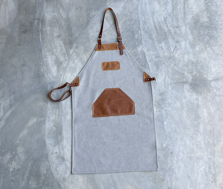 A light grey canvas apron with tan leather detaill and straps.  The updated apron design has crossover straps. Apron featured against a concrete screeded floor.