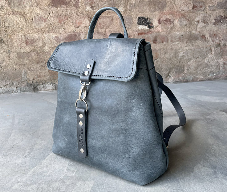 Denim blue leather backpack with black. Boasts secure pocket, key clip, inner pocket and power bank cable porthole. Closes securely with a metal clip.