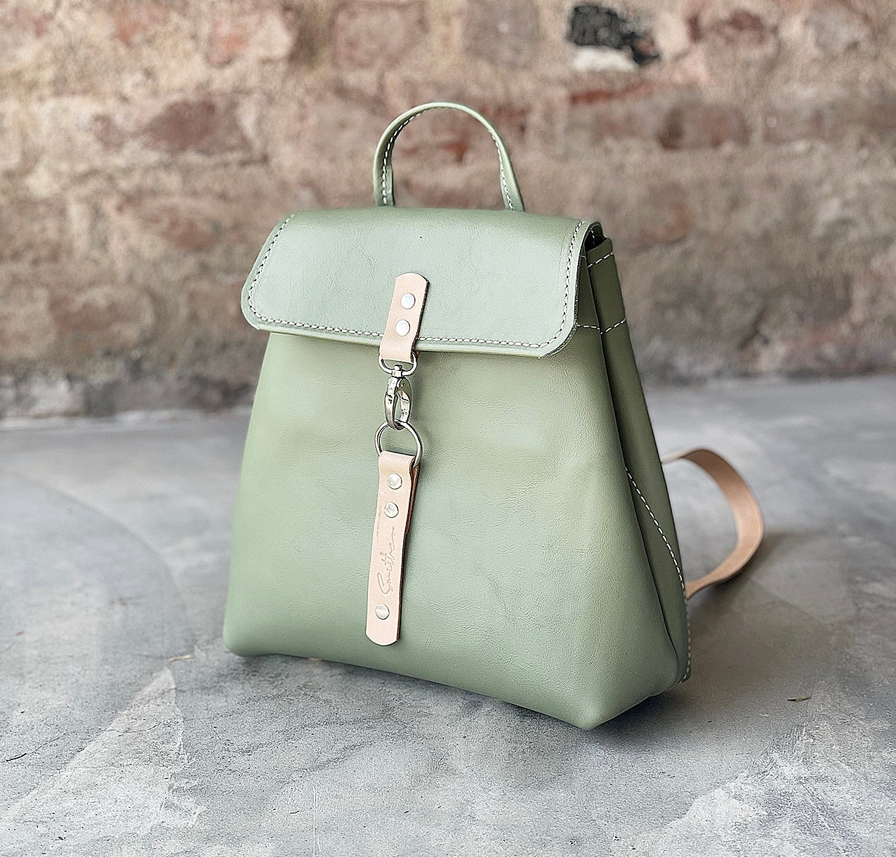 Green leather backpack with nude coloured detail. Boasts secure pocket, key clip, inner pocket and power bank cable porthole. Closes securely with a metal clip.