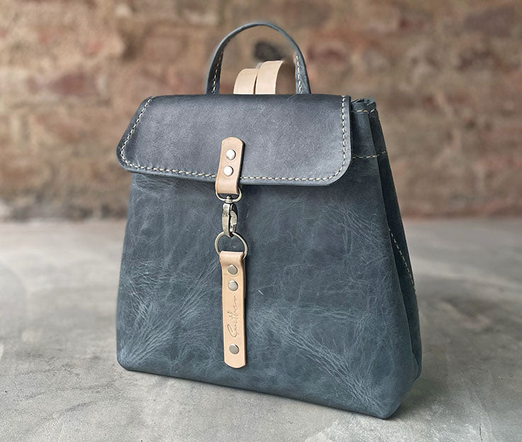 Denim blue leather backpack with nude colour detail. Boasts secure pocket, key clip, inner pocket and power bank cable porthole. Closes securely with a metal clip.