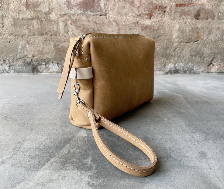 A fudge and nude colour combo leather toiletry bag featured against a red brick wall and concrete floor.  