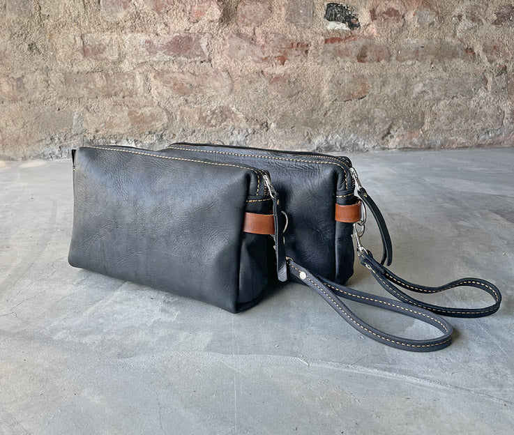 Two black and amber colour combo leather toiletry bags featured against a red brick wall and concrete floor.  