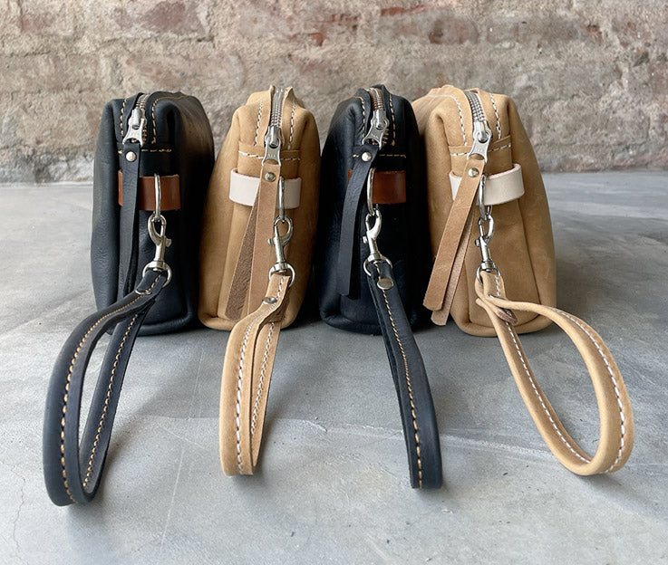 Leather toiletry bags in black and tan and fudge and nude colour detail against a rough red brick wall and concrete floor.